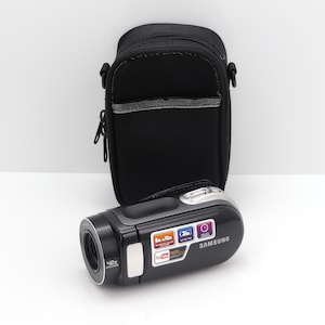 May include: A black Samsung camcorder with a 42x zoom lens and a silver top button, positioned in front of a black zippered carrying case. The camcorder features a rectangular label with text including 6hr to 15hr, 3hr battery time, and YouTube.
