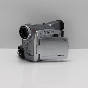 Puede incluir: Una videocámara digital Canon MV750i de color plateado con ranura para cinta Mini DV, lente de zoom óptico de 22x y pantalla LCD lateral abatible. El dispositivo incluye varios botones de control en la parte superior y el logotipo de Canon.