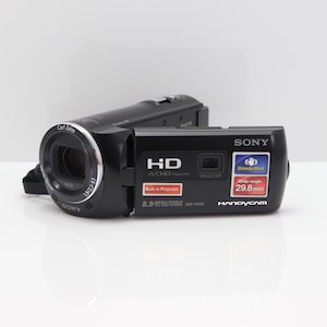 Op de afbeelding: Een zwarte Sony Handycam camcorder met een Carl Zeiss lens, voorzien van een open zijpaneel met een HD AVCHD logo, een ingebouwde projector label, 8.9 mega pixels tekst, SteadyShot branding en een groothoek 29.8mm lens specificatielabel.