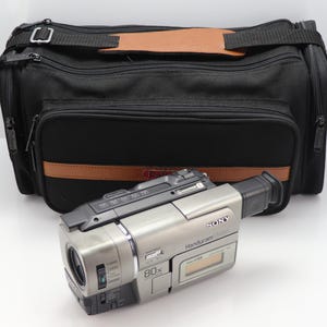 May include: A silver Sony Handycam video camera with an 80x zoom lens and a small LCD screen, positioned in front of a black rectangular carrying bag featuring a brown leather-like accent strip and a matching brown handle pad.