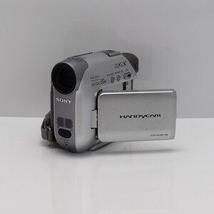May include: A silver Sony Handycam DCR-HC22E camcorder with a flip-out LCD screen. The device features a Carl Zeiss lens with 20x optical zoom, a built-in microphone, and various control buttons on the top and side of the compact camera body.