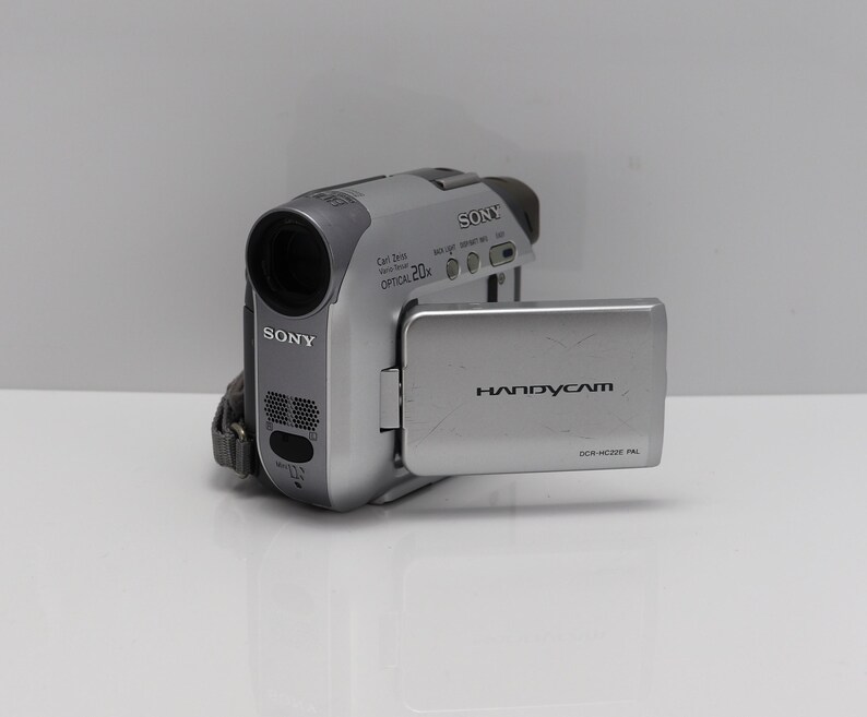 May include: A silver Sony Handycam DCR-HC22E camcorder with a flip-out LCD screen. The device features a Carl Zeiss lens with 20x optical zoom, a built-in microphone, and various control buttons on the top and side of the compact camera body.