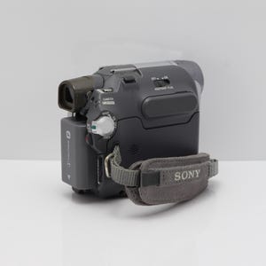 May include: A dark gray Sony camcorder featuring a rectangular battery pack, a viewfinder, and a gray fabric hand strap with the Sony logo printed in white. The device includes a mode dial, a NightShot Plus switch, and various control buttons.