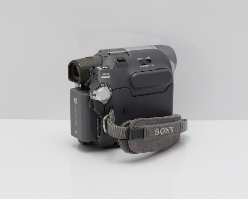May include: A dark gray Sony camcorder featuring a rectangular battery pack, a viewfinder, and a gray fabric hand strap with the Sony logo printed in white. The device includes a mode dial, a NightShot Plus switch, and various control buttons.
