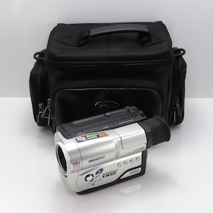 May include: A silver and black Samsung digital camcorder with a 22x zoom lens and 500x digital zoom capability, featuring an Easy-Q button and Digital Image Stabilizer, positioned in front of a black padded camera carrying bag with a top handle.