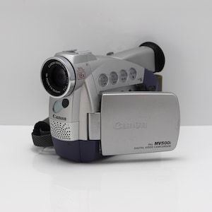 May include: A silver and dark blue Canon MV500i digital video camcorder with an 18x zoom lens, 360x digital zoom, and a flip-out LCD screen. The device features various control buttons on the top, a speaker grille, and the Canon brand name.