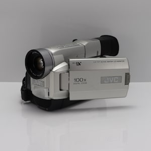 Pode incluir: Uma filmadora Mini DV JVC prateada com lente de zoom digital de 100x e monitor LCD de matriz ativa TFT de 2,5 polegadas. O dispositivo possui um visor preto na parte traseira e uma lente proeminente na frente, com o logotipo JVC na lateral.