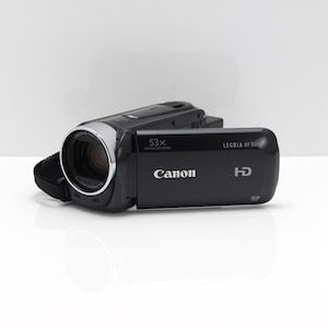 May include: A black Canon Legria HF R406 HD camcorder featuring a 53x advanced zoom lens. The device has a silver-rimmed lens, a side-hinged display panel with the Canon logo, and HD branding. A black fabric strap is attached to the side of the body.