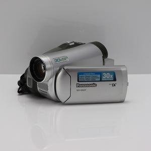 Op de afbeelding: Een zilverkleurige Panasonic NV-GS27 MiniDV-camcorder met een 30x optische zoomlens. Het zijpaneel bevat een blauwe sticker met de tekst Language Menu, met een lijst van diverse talen, en de 30x Optical Zoom-branding in witte en blauwe tekst.