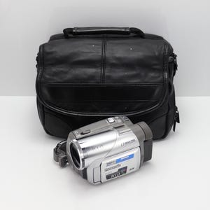 Puede incluir: Una videocámara Panasonic 3CCD plateada con lente, botones superiores y panel de pantalla lateral, situada frente a una bolsa de cámara de cuero negro con asa superior y cierre de solapa frontal.