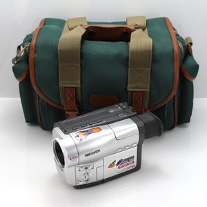 May include: A silver Samsung camcorder with a 440x digital zoom lens and various control buttons, positioned in front of a dark green canvas camera bag with tan webbing straps and brown leather-like trim accents on the edges and handle.