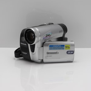 Op de afbeelding: Een zilveren en zwarte Panasonic NV-GS21 digitale camcorder met een 24x optische zoomlens. Het zijpaneel heeft een open scherm met een sticker met taalmenu-opties, een label voor ingebouwd LED-licht en een SD-kaartlogo.