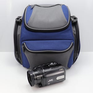 Puede incluir: Una mochila para cámara azul y gris con una videocámara JVC negra delante. La mochila tiene múltiples compartimentos con cremallera y un asa superior. La videocámara es negra con letras plateadas y una lente.