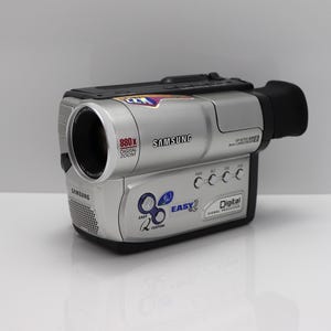 May include: A silver and black Samsung camcorder featuring an 880x digital zoom lens, a black viewfinder, and various control buttons on the side. The device displays labels including Easy-Q, Digital Signal Processor, and Samsung branding.