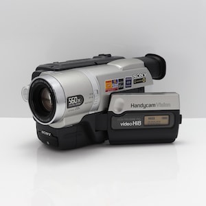 May include: A silver and black Sony Handycam Vision video Hi8 camcorder featuring a 560x digital zoom lens, a 20x optical zoom, and a side-mounted display screen with the text Handycam Vision and video Hi8 clearly visible on the device body.