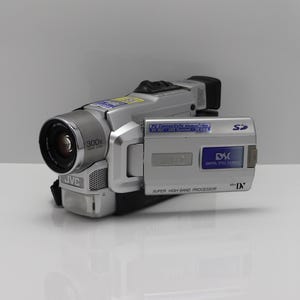 May include: A silver JVC MiniDV camcorder featuring a 300x digital zoom lens, a flip-out LCD screen with a Digital Still Camera logo, an SD card slot, and a Super High-Band Processor label. The device includes various blue informational stickers.