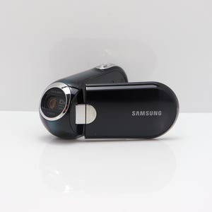 Peut inclure: Un caméscope numérique Samsung noir à la finition brillante, doté d'un écran latéral rotatif, d'un objectif cerclé d'argent avec l'inscription 10x optical zoom et d'une charnière argentée reliant le corps principal au panneau d'affichage.