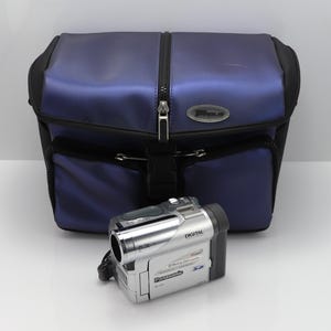 Puede incluir: Una videocámara digital Panasonic plateada con compatibilidad para tarjetas de memoria SD, situada frente a un estuche de transporte con cremallera en color azul oscuro y negro. La cámara tiene un objetivo, un botón superior y marca.