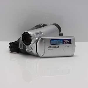 Op de afbeelding: Een zilverkleurige Panasonic NV-GS60 MiniDV-camcorder met een 30x optische zoomlens. Het apparaat heeft een uitklapbaar scherm met een taalmenu en de tekst 30x Optical Zoom. De behuizing is zilverkleurig met zwarte accenten en een zwarte polsband.