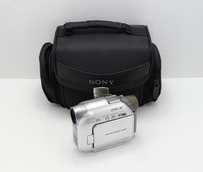 May include: A silver Sony Handycam digital video camera with a 20x optical zoom lens, positioned in front of a black Sony branded carrying case. The camera features a flip-out screen, multiple control buttons, and clear text labels on the body.