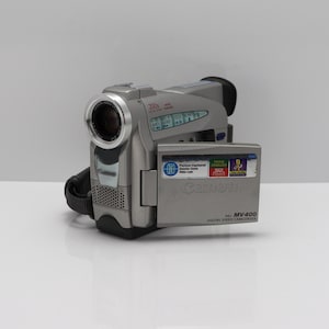 May include: A silver Canon MV400 digital video camcorder with a flip-out LCD screen. The device features a 200x digital zoom lens, a black hand strap, and labels indicating MiniDV and PAL compatibility. The Canon logo is visible on the body.