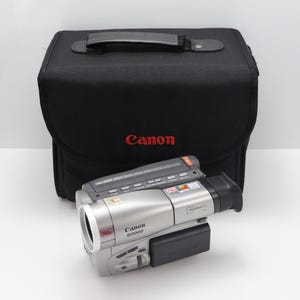 Op de afbeelding: Een zilverkleurige Canon G2000 camcorder met een bedieningspaneel aan de bovenzijde en een zwarte accu, geplaatst voor een zwarte Canon draagtas. De camcorder is voorzien van diverse knoppen, een lens en een zoeker aan de rechterkant.