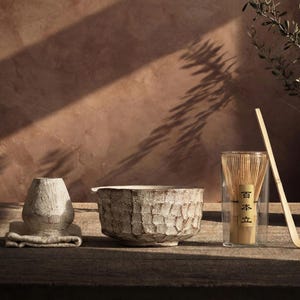 May include: A still-life arrangement featuring a textured ceramic bowl, a vase, and a bamboo whisk. The bowl and vase have a woven pattern. A bamboo whisk is in a glass container. The items are on a wooden surface.