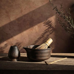 Puede incluir: Un juego de té matcha con un cuenco de cerámica marrón oscuro, un batidor de bambú, un pequeño recipiente de cerámica y una cuchara de bambú. El juego está dispuesto sobre una superficie de madera con un fondo cálido y terroso.