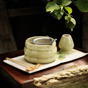 May include: A matcha tea set featuring a light green ceramic bowl with a stainless steel strainer, a whisk, and a bamboo scoop. The set is arranged on a rectangular tray with floral accents, perfect for preparing matcha tea.