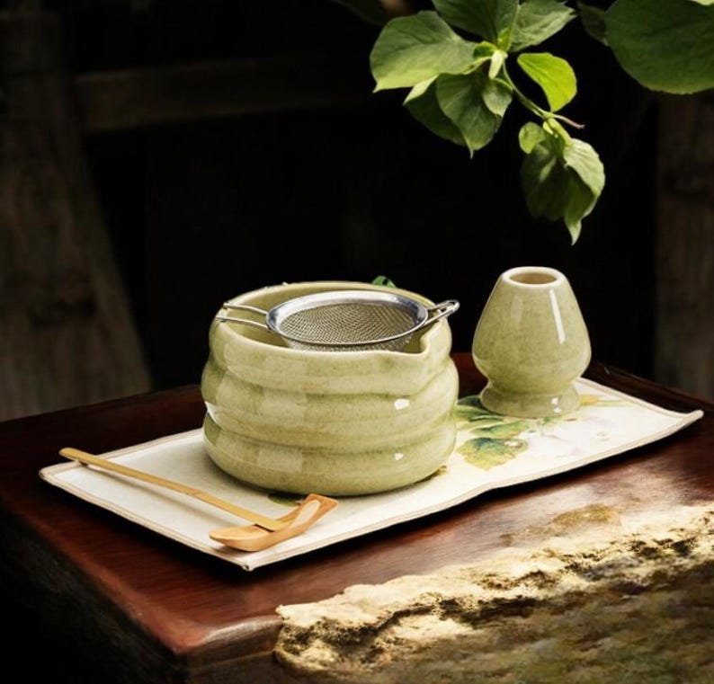 May include: A matcha tea set featuring a light green ceramic bowl with a stainless steel strainer, a whisk, and a bamboo scoop. The set is arranged on a rectangular tray with floral accents, perfect for preparing matcha tea.