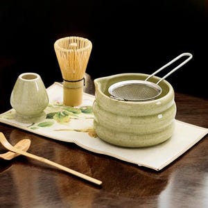 May include: A matcha tea set featuring a green ceramic bowl with a stainless steel strainer, a bamboo whisk, a small ceramic container, and a bamboo scoop. The set is arranged on a floral patterned mat.