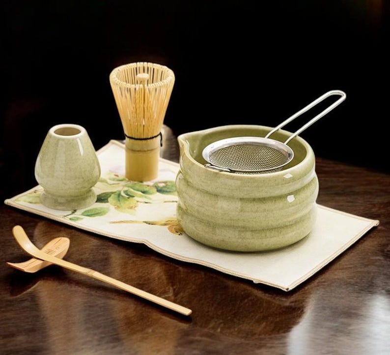 May include: A matcha tea set featuring a green ceramic bowl with a stainless steel strainer, a bamboo whisk, a small ceramic container, and a bamboo scoop. The set is arranged on a floral patterned mat.