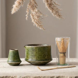 Könnte beinhalten: Ein Matcha-Teeset mit einer grünen Keramikschale, einer kleinen grünen Keramikvase, einem Bambusbesen in einem transparenten Behälter und einem Bambuslöffel. Das Set wird auf einer neutralen Oberfläche mit getrocknetem Ziergras im Hintergrund präsentiert.