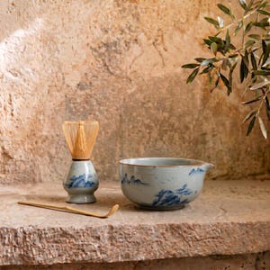 Puede incluir: Un juego de té matcha con un cuenco de cerámica con boquilla, un batidor y una cuchara. El cuenco y la base del batidor están decorados con diseños de paisajes azules. El batidor es de bambú.