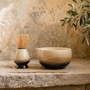 Puede incluir: Un juego de té matcha con un cuenco de cerámica beige y marrón, un batidor de bambú y una cuchara de bambú. El juego se exhibe sobre una superficie de piedra, con ramas de olivo en el fondo.