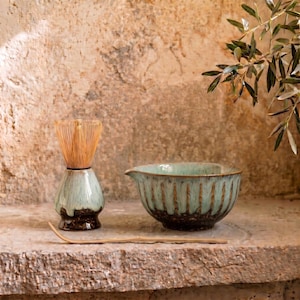 May include: A matcha tea set featuring a ceramic bowl, whisk, and whisk holder. The bowl is teal and brown, and the whisk is made of bamboo. The set is displayed on a stone surface.