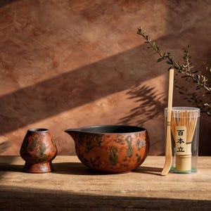 May include: A ceramic matcha tea set with a brown and green speckled pattern. The set includes a bowl, a small vase, a bamboo whisk, and a scoop. The whisk is in a clear container with Japanese text.