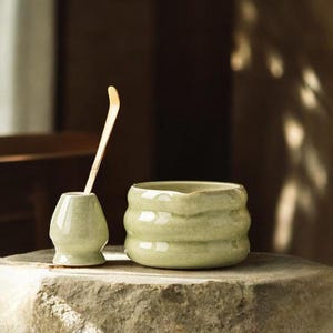 May include: A light green ceramic tea bowl and whisk holder with a bamboo whisk. The tea bowl has a textured, ribbed design. The objects are displayed on a stone surface, suggesting a tea ceremony setting.