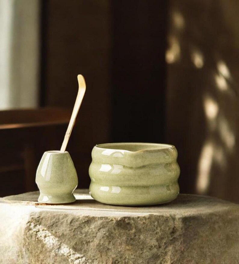 May include: A light green ceramic tea bowl and whisk holder with a bamboo whisk. The tea bowl has a textured, ribbed design. The objects are displayed on a stone surface, suggesting a tea ceremony setting.