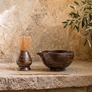 Puede incluir: Un cuenco de matcha de cerámica marrón con un pico, un batidor de bambú y una cuchara de bambú. El cuenco y el soporte del batidor tienen un acabado texturizado y esmaltado. Los artículos están colocados sobre una superficie de piedra.