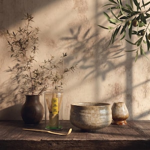 Puede incluir: Un juego de té matcha que incluye un jarrón de cerámica marrón, un batidor de bambú dentro de un estuche cilíndrico transparente con una etiqueta amarilla que dice 百本立, un cuenco de cerámica beige moteado, un soporte para batidor marrón y una cuchara de té de bambú curva.