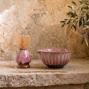 May include: A tea ceremony set featuring a pink and brown ceramic bowl with a spout, a matching whisk holder, a bamboo whisk, and a bamboo spoon. The set is displayed on a stone surface with a neutral background.