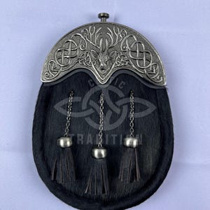 Puede incluir: Sporran de cuero negro con una cabeza de ciervo plateada y un diseño de nudo celta en la solapa superior. El sporran tiene tres borlas negras.