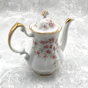 Peut inclure: Une théière en porcelaine blanche avec un motif floral et des finitions dorées. La théière a un couvercle et un bec verseur. La théière est posée sur un fond blanc.