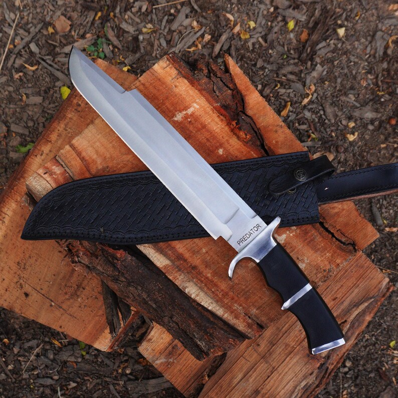 May include: A silver Predator brand knife with a black handle and a black leather sheath. The knife is resting on a piece of wood.