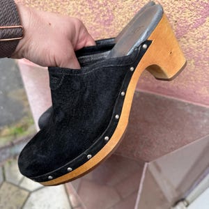 Könnte beinhalten: Schwarze Clogs aus Wildleder mit Holzabsatz und silberfarbenen Nieten. Die Clogs haben eine runde Spitze und ein Slip-on-Design. Der Absatz ist etwa 7,6 cm hoch. Der Markenname ist auf der Innensohle sichtbar.
