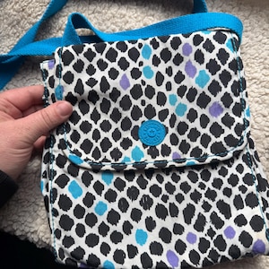 May include: A small, square crossbody bag with a flap closure. The bag features a white background with a black, dalmatian-like pattern, accented with blue and purple spots. It has a bright blue strap and a matching blue button closure.