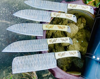 May include: Five kitchen knives with patterned blades and white handles with floral designs. The knives are arranged in a row, with the largest knife at the bottom and the smallest knife at the top. The knives are resting on a brown wooden surface.