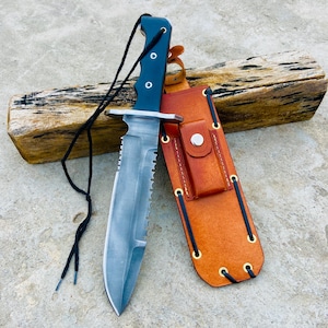 May include: A survival knife with a serrated blade and a black handle. The knife is paired with a brown leather sheath. A black cord is attached to the handle. The knife is resting on a piece of wood.