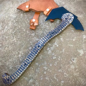 May include: A blue and white patterned axe with a dark blue blade and a brown leather sheath. The axe handle has a repeating oval design. The sheath has a strap with a snap closure. The axe is approximately 15 inches long.